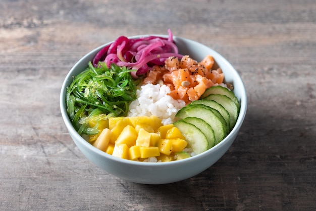 Poke bol avec riz saumonconcombremangueoignonsalade wakame graines de pavot et graines de tournesol sur table en bois