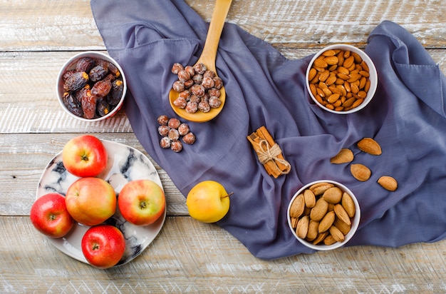 Pommes dans une assiette avec des bâtons de cannelle, des dates, des amandes pelées et non pelées dans des bols, des noix dans une vue de dessus de cuillère en bois sur bois et textile