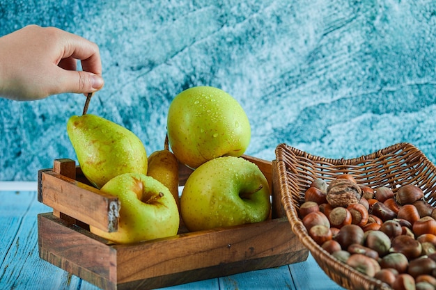 Pommes et poire dans un panier en bois et bol de noisettes sur table bleue.