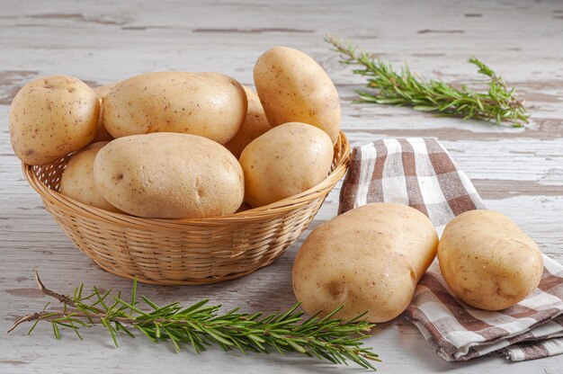 Pommes de terre crues dans un panier en osier tressé avec des feuilles de romarin naturel sur une table rustique en bois