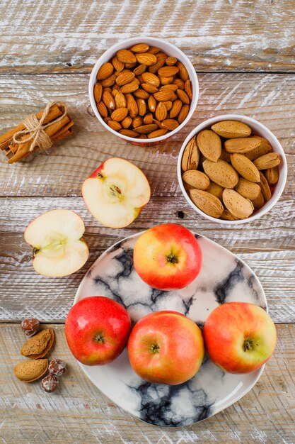 Pommes et tranches en plaque avec des bâtons de cannelle, amandes pelées et non pelées dans des bols, noix vue de dessus sur un bois