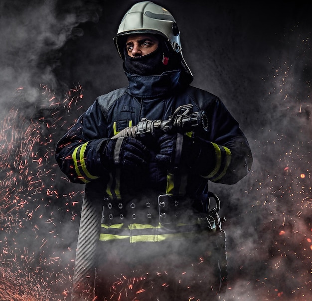 Un pompier professionnel en uniforme tient le tuyau d'incendie dans des étincelles de feu et de la fumée sur un fond sombre.