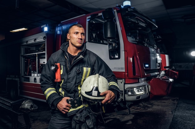 Pompier en uniforme tenant un casque et regardant une caméra tout en se tenant près d'un camion de pompiers dans un garage d'un service d'incendie la nuit.