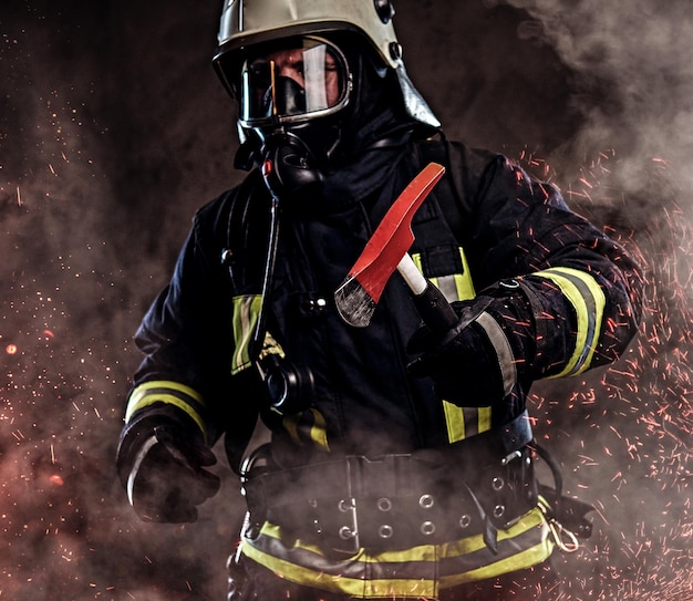 Un pompier vêtu d'un uniforme et d'un masque à oxygène tient une hache rouge debout dans des étincelles de feu et de la fumée sur un fond sombre.