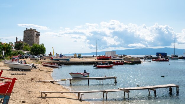 Port de mer, bateaux amarrés sur la mer Égée, quelques voitures en stationnement, deux petites jetées en bois et Tour de Prosphorion, Ouranoupolis, Grèce