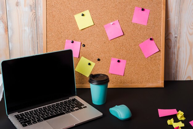 Portable; tasse de café à emporter; souris et liège avec notes autocollantes sur le bureau noir