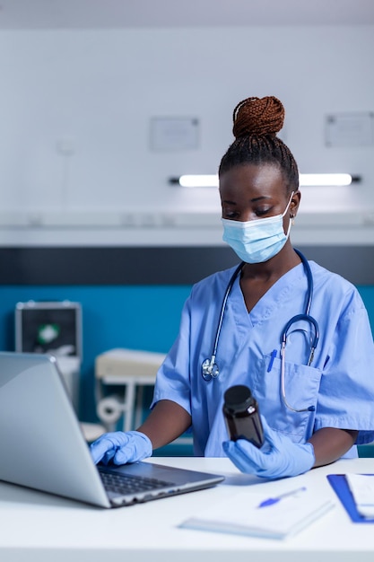 Porter un masque facial à l'aide d'un ordinateur moderne pour lire le prospectus antiviral prescrit. Infirmière de clinique tenant une bouteille de médicament vérifiant la date de péremption et les ingrédients du médicament.