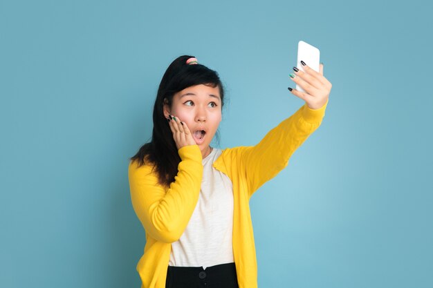 Portrait d'adolescent asiatique isolé sur fond bleu studio. Beau modèle femme brune aux cheveux longs. Concept d'émotions humaines, expression faciale, ventes, publicité. Faire un selfie ou un vlog.