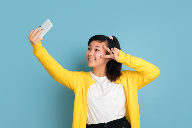 Portrait d'adolescent asiatique isolé sur fond bleu studio. Beau modèle femme brune aux cheveux longs. Concept d'émotions humaines, expression faciale, ventes, publicité. Faire un selfie ou un vlog.