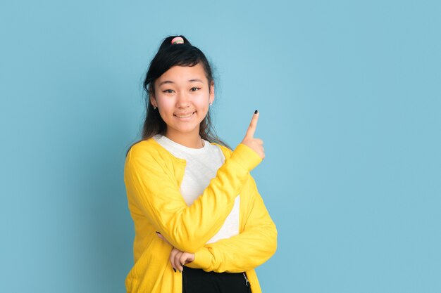 Portrait d'adolescent asiatique isolé sur fond bleu studio. Beau modèle femme brune aux cheveux longs. Concept d'émotions humaines, expression faciale, ventes, publicité. Souriant, pointant vers le haut, a l'air mignon.