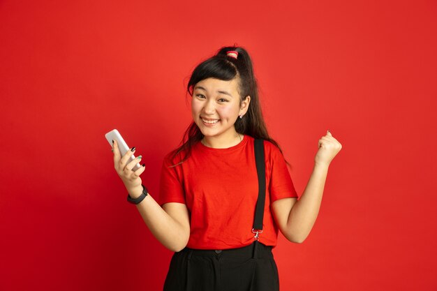 Portrait d'adolescent asiatique isolé sur fond de studio rouge. Beau modèle brune féminine dans un style décontracté. Concept d'émotions humaines, expression faciale, ventes, publicité. Heureux, tenant le smartphone.