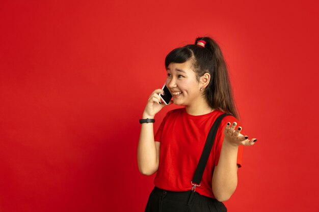 Portrait d'adolescent asiatique isolé sur fond de studio rouge. Beau modèle femme brune aux cheveux longs dans un style décontracté. Concept d'émotions humaines, expression faciale, ventes, publicité. Parler au téléphone.