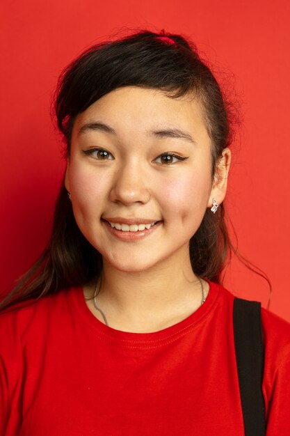 Portrait d'adolescent asiatique isolé sur fond de studio rouge. Beau modèle femme brune aux cheveux longs dans un style décontracté. Concept d'émotions humaines, expression faciale, ventes, publicité. Sourire mignon.