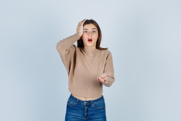 Portrait d'une adolescente gardant la main sur la tête, étirant la main à l'avant dans un pull, un jean et à la vue de face choquée