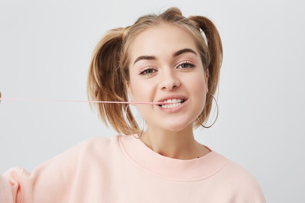 Portrait d'une adolescente joyeuse et joyeuse avec une peau propre et saine, des yeux noirs, deux queues de cheval en pull rose posant sur fond gris, montrant ses dents blanches et étirant le chewing-gum