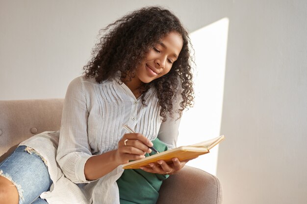 Portrait d'adolescente mignonne d'origine africaine assise confortablement sur un canapé avec un cahier, faisant des dessins ou des croquis, ayant inspiré un regard joyeux. Élégante jeune femme noire écrit dans son journal
