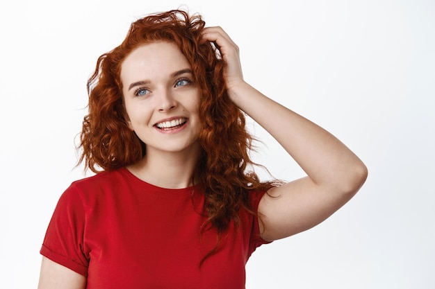 Portrait d'une adolescente mignonne et rêveuse aux cheveux bouclés rouges, souriante et regardant de côté l'espace de copie gauche, touchant une coupe de cheveux saine et naturelle au gingembre sur blanc