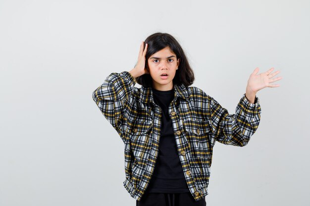 Portrait d'une adolescente tenant la main sur la tête, montrant un geste de bienvenue en chemise décontractée et regardant la vue de face choquée