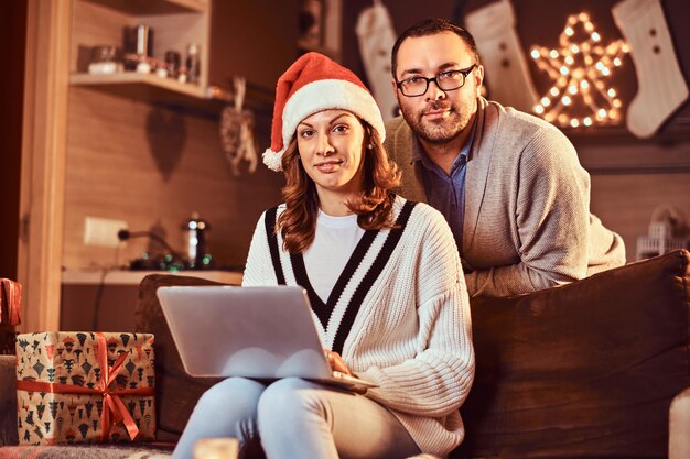 Portrait d'un adorable couple avec ordinateur portable regardant la caméra célébrant le réveillon de Noël à la maison.