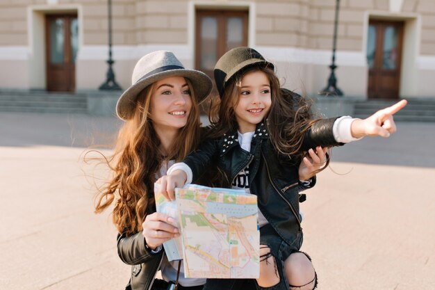 Portrait d'adorable petite fille au chapeau à la mode pointant avec le doigt sur les sites touristiques de la nouvelle ville pendant le voyage avec maman Charmante femme portant une fille joyeuse tenant une carte et regardant autour avec le sourire.