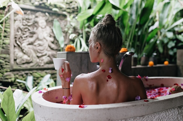 Portrait de l'arrière d'une femme européenne gracieuse prenant un bain avec des pétales de rose et une dégustation de thé.