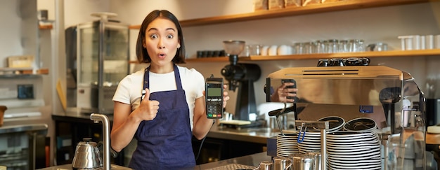 Photo gratuite portrait d'une barista de café montrant le pouce levé et un lecteur de cartes recommandant une machine à payer sans contact