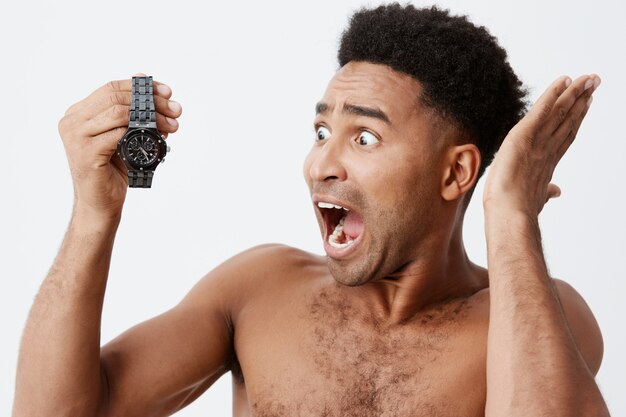 Portrait de beau beau mâle afro-américain aux cheveux bouclés sans vêtements tenant la montre à la main dans les mains, regardant avec une expression stressée et confuse, criant fort