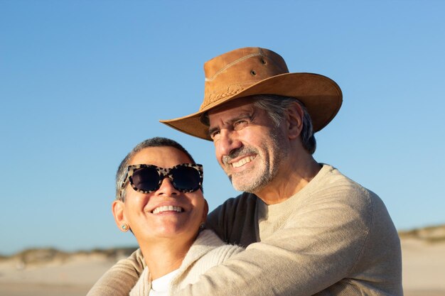 Portrait d'un beau couple de personnes âgées qui passe du temps ensemble à l'extérieur par une journée ensoleillée. Bel homme aux cheveux gris souriant en chapeau de cow-boy étreignant heureuse dame aux cheveux courts en lunettes de soleil. Amour, concept de loisirs