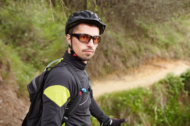 Portrait de beau cycliste professionnel confiant en vêtements de sport, lunettes, casque et sac à dos