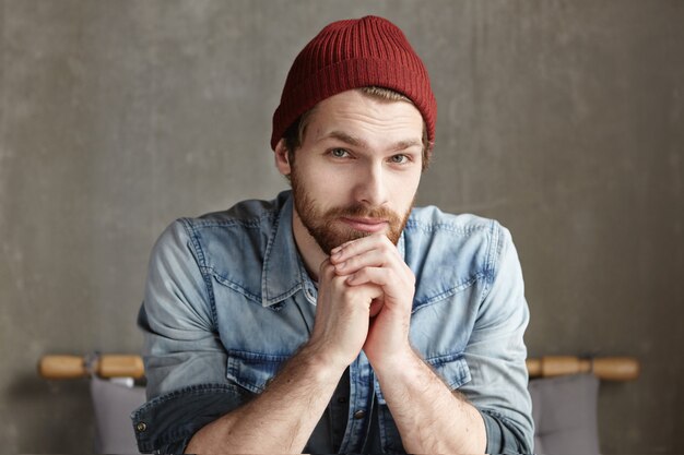 Portrait de beau hipster barbu avec un sourire amical et des yeux gentils assis seul dans l'intérieur d'un café moderne, en attente d'amis contre un mur de béton gris