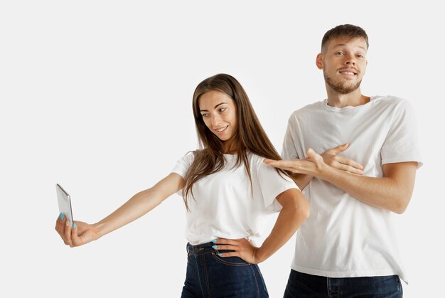 Portrait de beau jeune couple isolé sur mur blanc. Expression faciale, émotions humaines, concept publicitaire. Femme faisant selfie ou vidéo pour vlog, l'homme s'ennuie, ne veut pas faire.