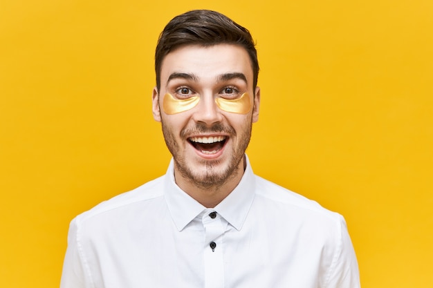 Portrait de beau jeune employé de bureau masculin énergique ayant une expression faciale excitée portant des patchs oculaires pour réduire les poches, les signes de fatigue et de stress, en gardant la bouche grande ouverte