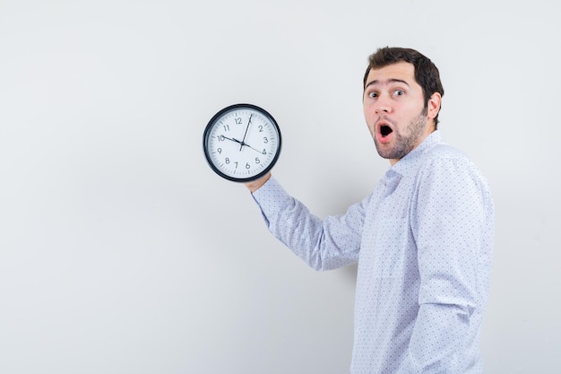 Photo gratuite portrait de beau jeune homme en chemise à motifs blancs montrant l'horloge et inquiet du temps isolé sur fond blanc