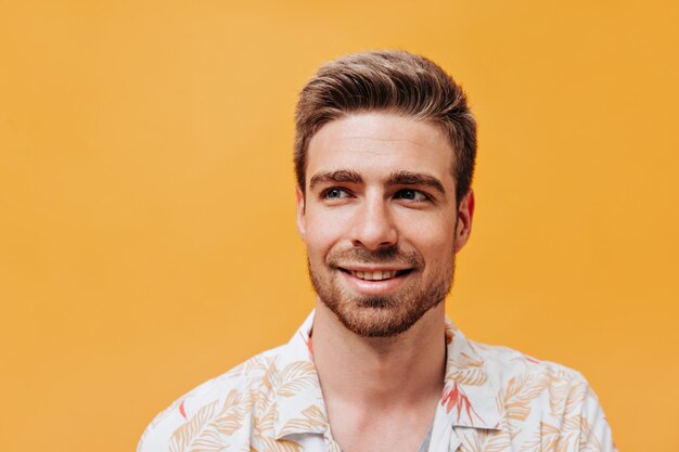 Portrait d'un beau jeune homme avec une coiffure élégante, des yeux bleus et une barbe en chemise légère et fraîche regardant loin et souriant sur un mur orange