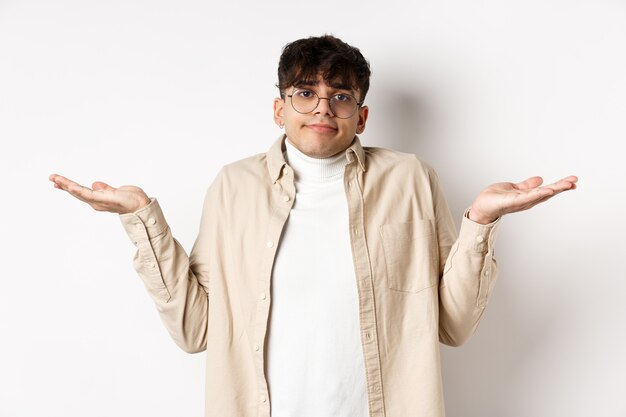 Portrait d'un beau jeune homme qui a l'air confus, ne sait rien, haussant les épaules et écartant les mains sur le côté sans aucune idée, debout sur fond blanc