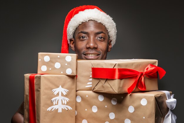 Le portrait de beau jeune homme souriant noir en bonnet de Noel avec des cadeaux sur l'obscurité.