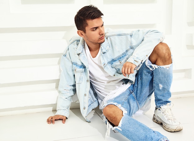 Portrait de beau jeune mannequin homme habillé en jeans assis près d'un mur texturé blanc