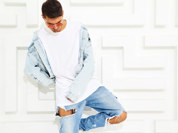 Portrait de beau jeune mannequin homme habillé en jeans posant près du mur texturé blanc