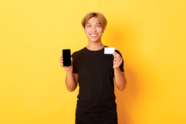 Portrait de beau jeune mec asiatique montrant l'écran du smartphone, application bancaire et carte de crédit, mur jaune debout et souriant.