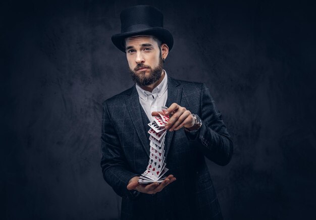 Portrait d'un beau magicien en costume noir, montrant un tour avec des cartes à jouer sur fond sombre.