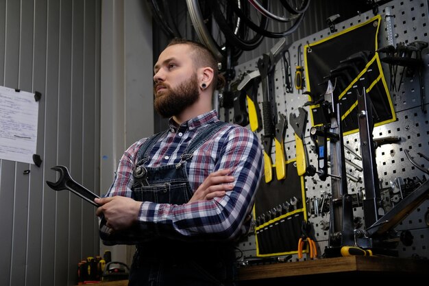 Portrait d'un beau mâle élégant avec barbe et coupe de cheveux portant une chemise en flanelle et une combinaison de jeans, tient une clé en acier, debout dans un atelier contre des outils muraux.