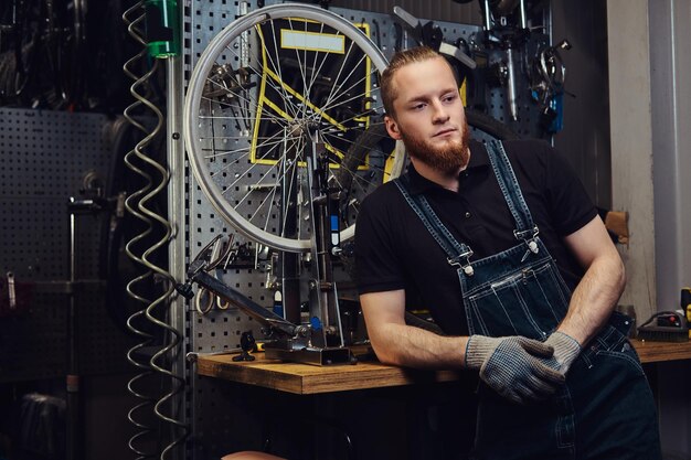 Portrait d'un beau mâle rousse avec barbe et coupe de cheveux portant une combinaison de jeans, debout près d'une roue de bicyclette dans un atelier.