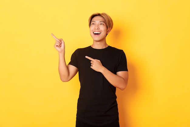 Portrait de beau mec asiatique heureux en t-shirt noir, pointant du doigt et regardant le coin supérieur gauche avec sourire satisfait, mur jaune