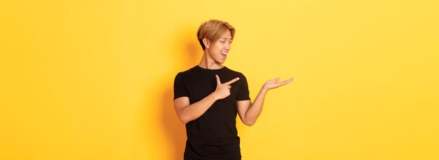 Portrait d'un beau mec asiatique souriant tenant quelque chose à portée de main et pointant le doigt dessus debout