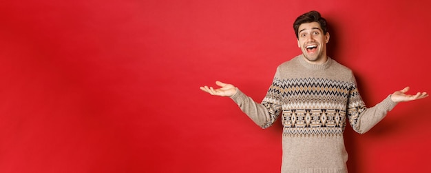 Portrait d'un beau mec caucasien heureux et surpris, portant un pull de Noël, écartant les mains sur le côté et l'air désemparé, debout sur fond rouge.