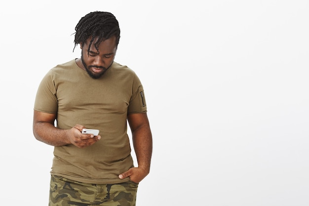 Portrait de beau mec dans un t-shirt marron posant contre le mur blanc avec son téléphone