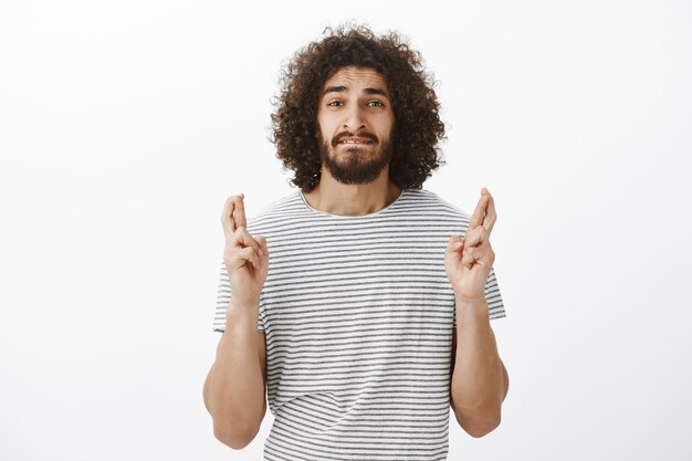 Portrait de beau mec hispanique inquiet avec une coiffure afro en t-shirt rayé, mordant la lèvre anxieusement et croisant les doigts dans l'espoir ou en priant, souhaitant que le rêve devienne réalité