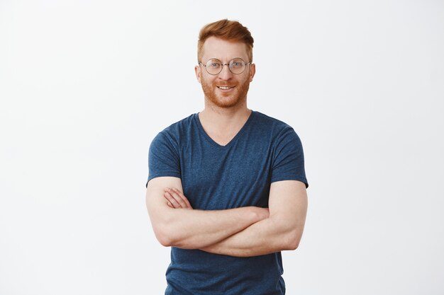 Portrait de beau mec mature rousse confiant avec poils dans des verres ronds, tenant les mains sur la poitrine et souriant largement