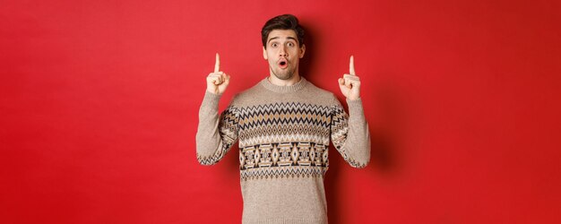 Portrait d'un beau mec surpris et étonné montrant une offre promotionnelle de noël, portant un pull de noël et debout sur fond rouge