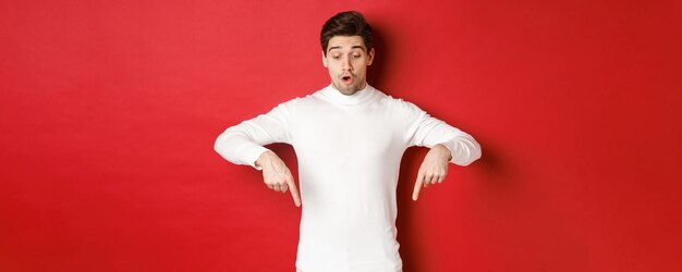 Portrait d'un beau mec surpris en pull blanc, regardant et pointant les doigts vers le logo, debout sur fond rouge
